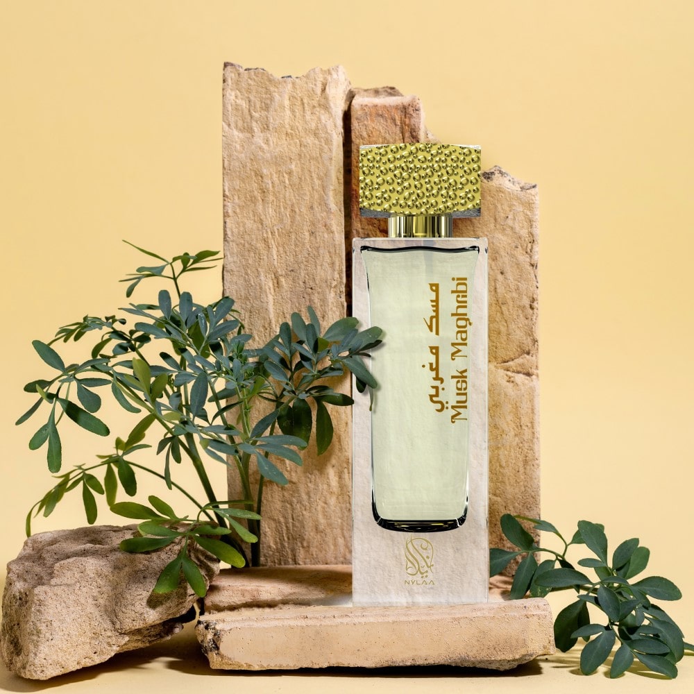 A transparent white perfume bottle by from Nylaa by Savia Exclusive with a golden cap across light brown wood & green leaves