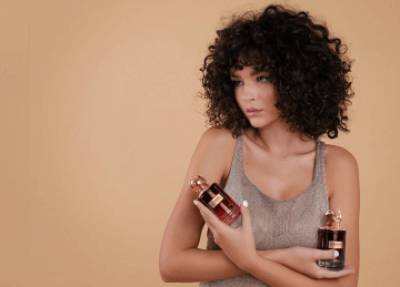 A women with curly hair wearing a silver dress holding two perfumes by Savia Exclusive